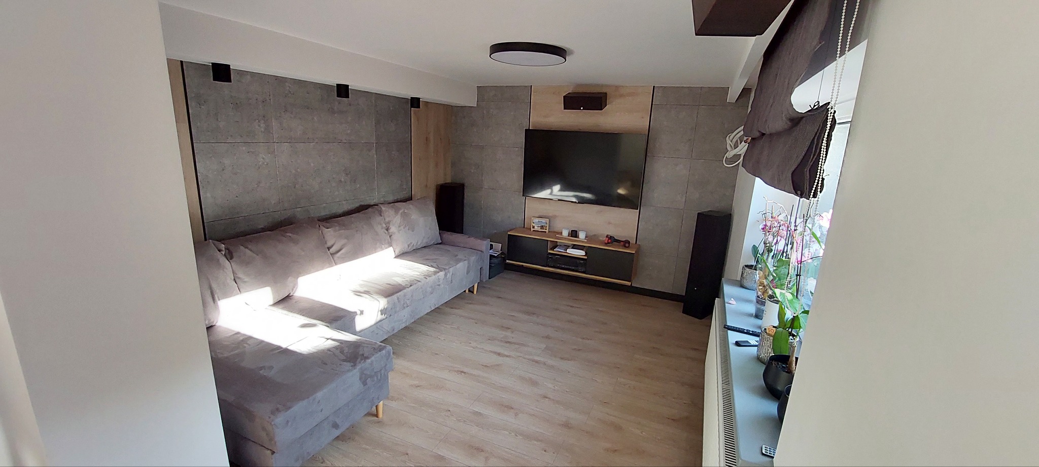 Szary narożnik z poduszkami w salonie z szarą ścianą z betonowych płyt, drewnianą podłogą i czarnym telewizorem na ścianie.