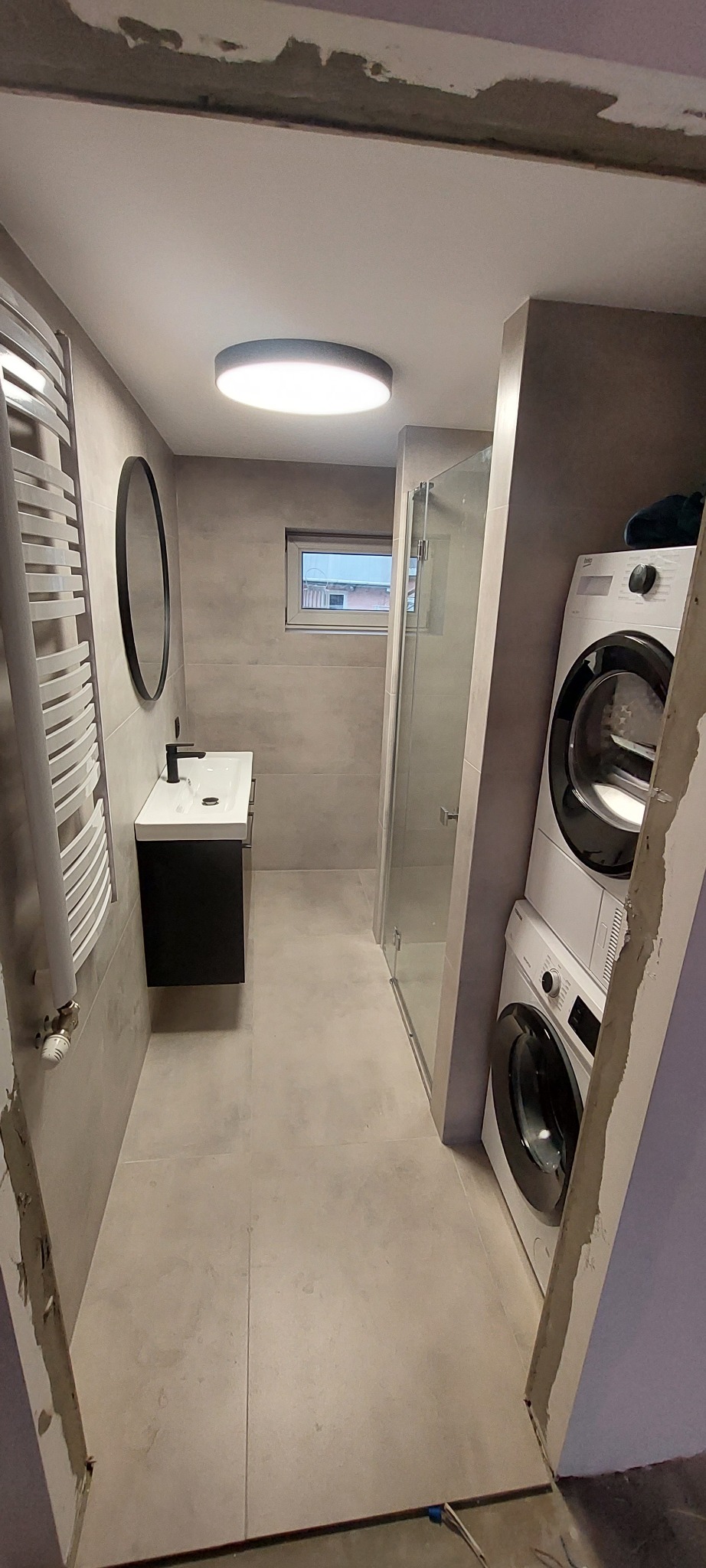 Wnętrze łazienki w szarych tonacjach z umywalką, okrągłym lustrem, grzejnikiem drabinkowym, kabiną prysznicową i pralko-suszarką ustawioną w pionie, widoczne niedokończone wykończenia przy futrynie.