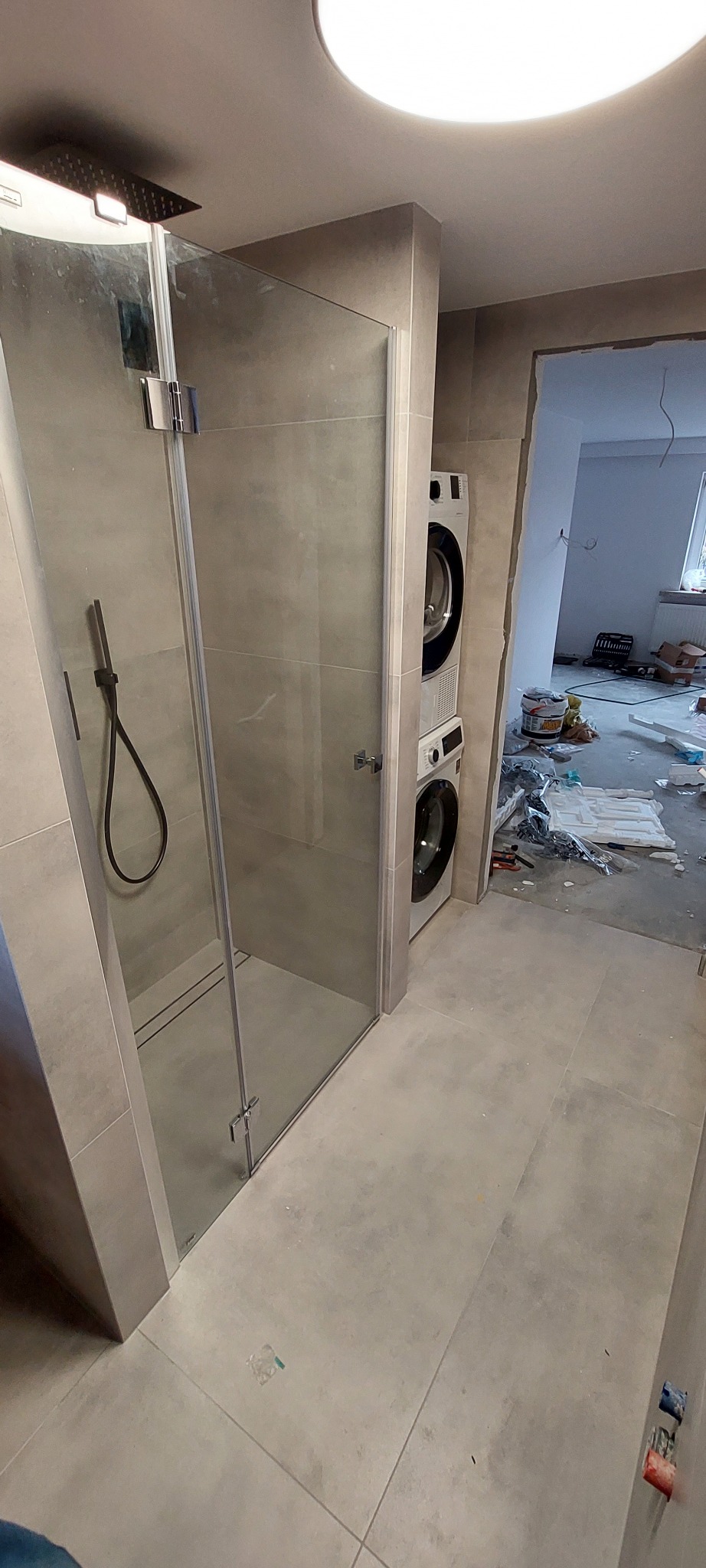 Szklana kabina prysznicowa z deszczownicą w łazience w trakcie prac, obok pralko-suszarka w zabudowie i widoczny bałagan remontowy w przejściu.
