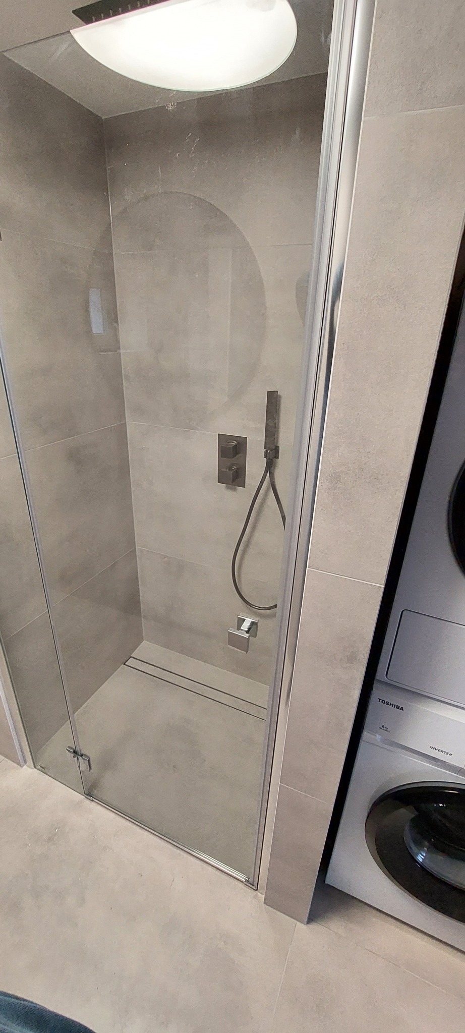Szklana kabina prysznicowa z szarymi płytkami, zamontowana bateria podtynkowa z deszczownicą i słuchawką, obok pralka Toshiba.