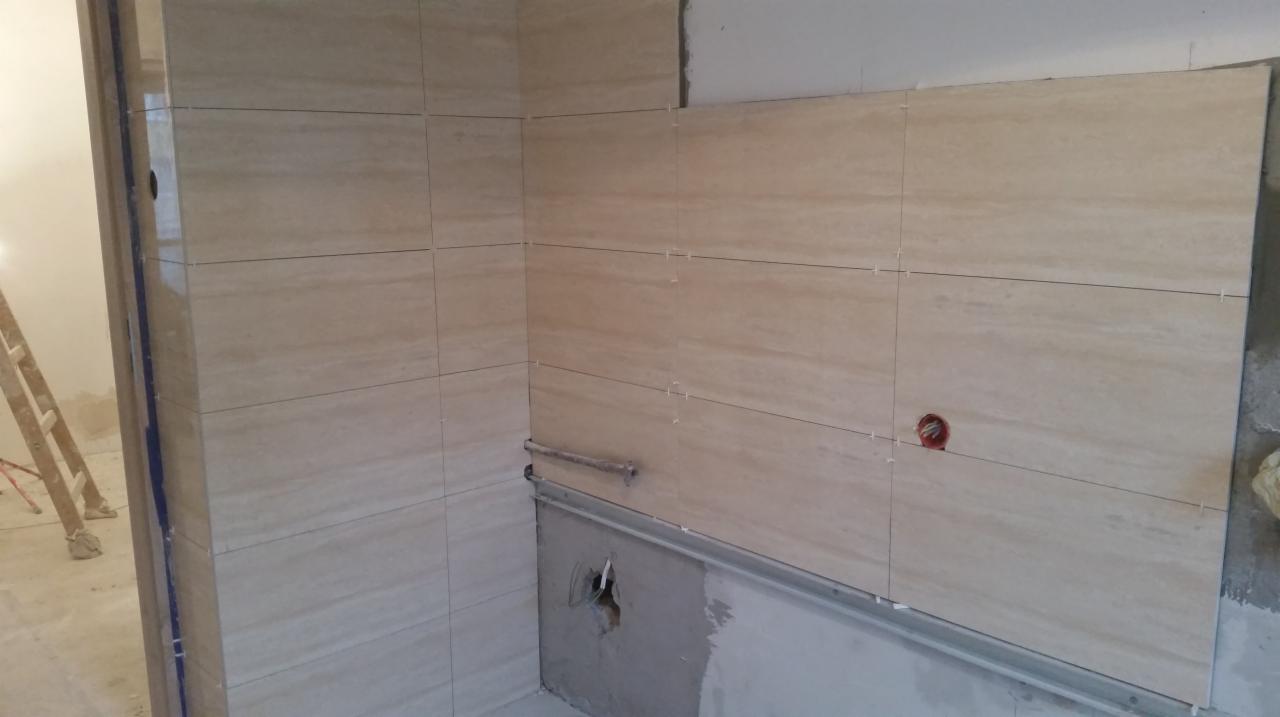 Ściana w trakcie układania płytek imitujących trawertyn w łazience, widoczne listwy montażowe i krzyżyki dystansowe, częściowo wykończone narożniki i przygotowane miejsce pod instalację wodną.