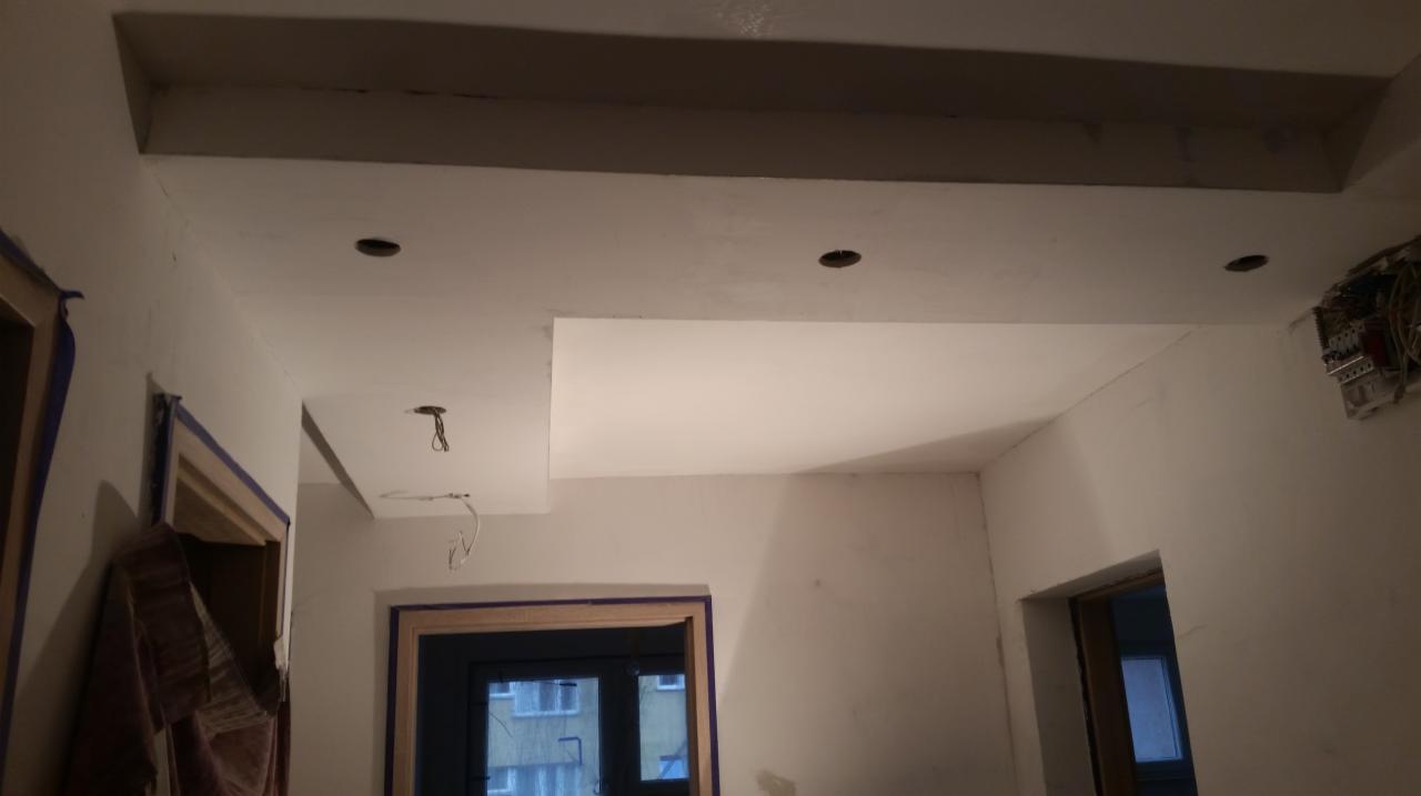 Nowoczesny sufit podwieszany z oświetleniem punktowym w jasnym wnętrzu. Widoczne przygotowane otwory na lampy i wyprowadzone kable. Wykończenie ścian w jasnym odcieniu.