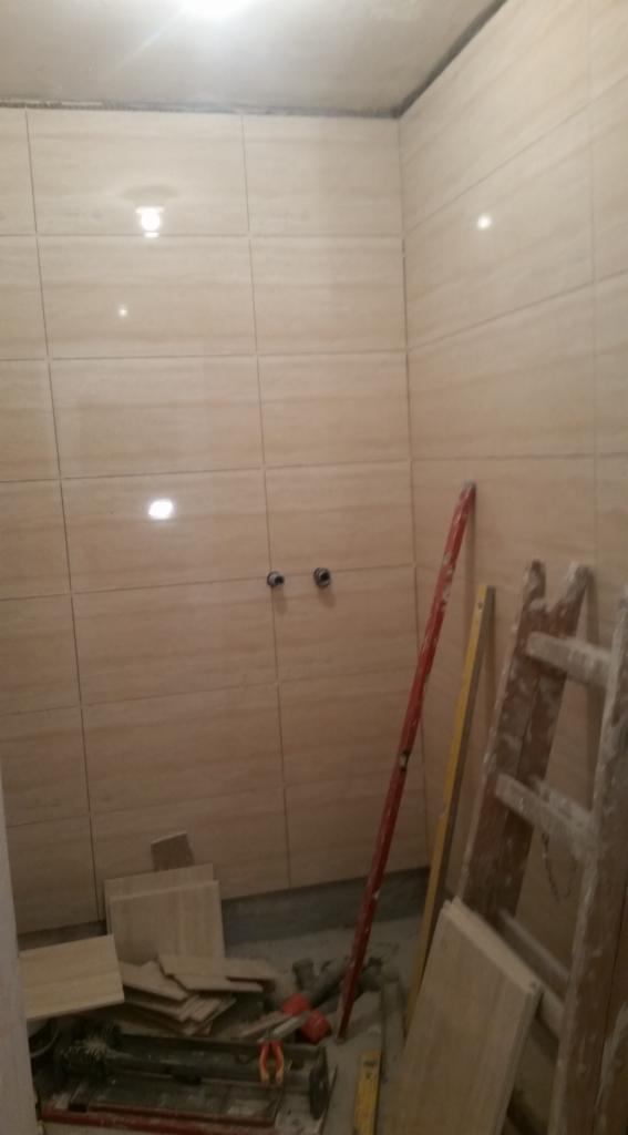 Ściany łazienki wyłożone beżowymi płytkami imitującymi drewno, widoczne rury, poziomica oraz drabina malarska, sugerujące etap prac wykończeniowych.