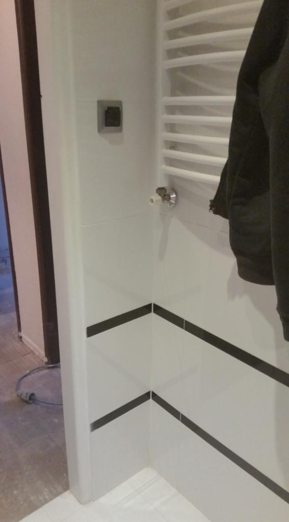 Wykończona łazienka z białymi płytkami ozdobionymi czarnymi paskami, grzejnik drabinkowy na ścianie, widoczny fragment ubrania i gniazdko elektryczne.