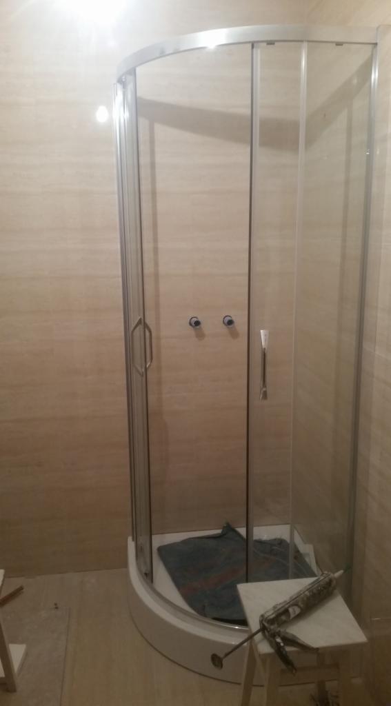 Narożna kabina prysznicowa z przezroczystego szkła z chromowanymi okuciami, zamontowana w łazience wyłożonej beżowymi płytkami imitującymi trawertyn, widoczny pistolet do silikonu na stołku.