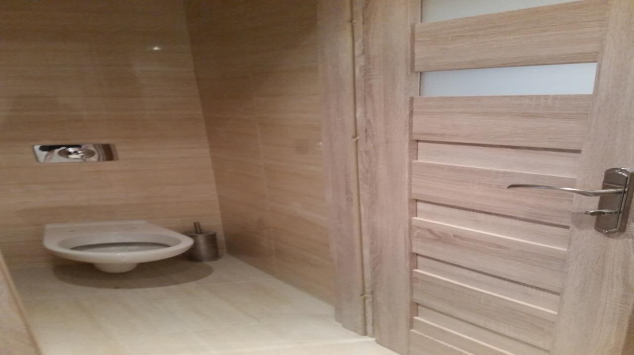 Nowoczesna łazienka z beżowymi płytkami imitującymi drewno, wisząca toaleta z chromowanym przyciskiem spłukiwania i drewniane drzwi z poziomymi panelami.