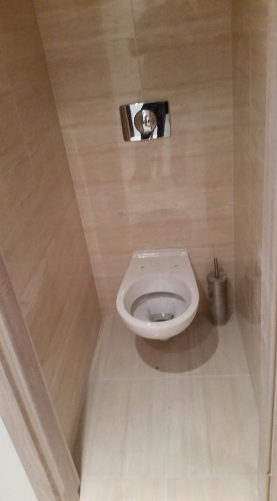 Wisząca muszla klozetowa zamontowana na ścianie wyłożonej płytkami imitującymi trawertyn, z widocznym chromowanym przyciskiem spłuczki i szczotką do WC.