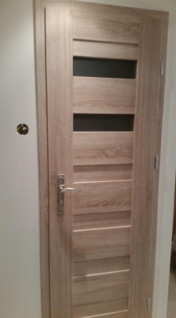 Zamknięte drzwi wewnętrzne z drewnopodobnym fornirem, srebrną klamką i trzema poziomymi szybami z czarnego szkła, widoczne na tle białej ściany z otworem na gniazdko elektryczne.