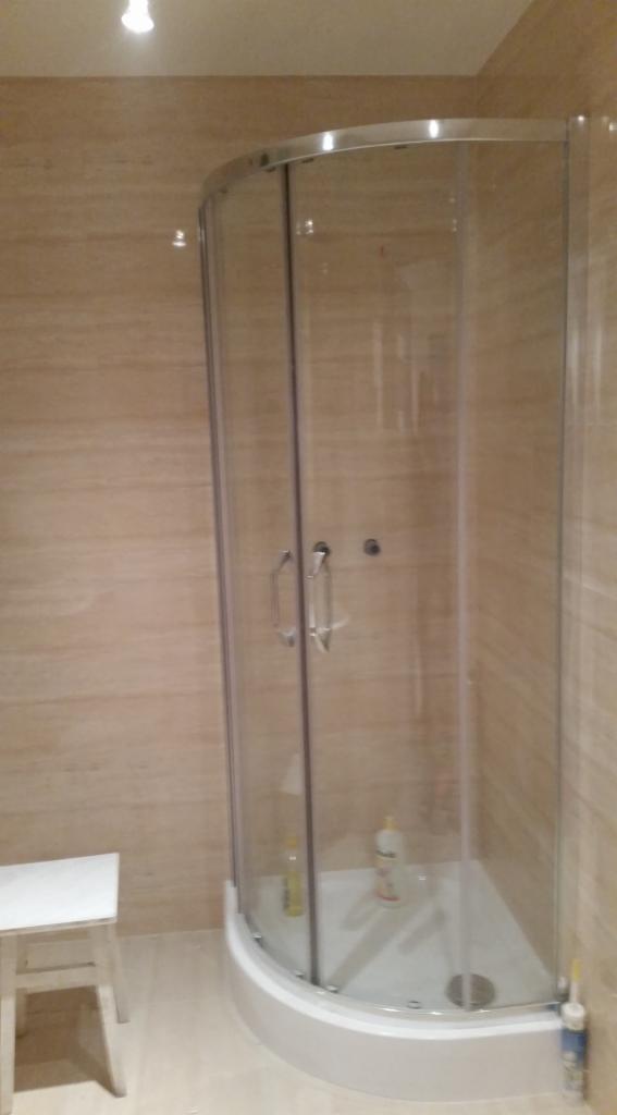 Narożna kabina prysznicowa z przezroczystymi drzwiami i srebrnymi uchwytami, biały brodzik, beżowe płytki na ścianach, biały stołek w rogu.