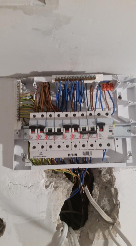 Otwarta skrzynka rozdzielcza z zainstalowanymi bezpiecznikami marki EATON i podłączonymi przewodami w ścianie z uszkodzonym tynkiem.