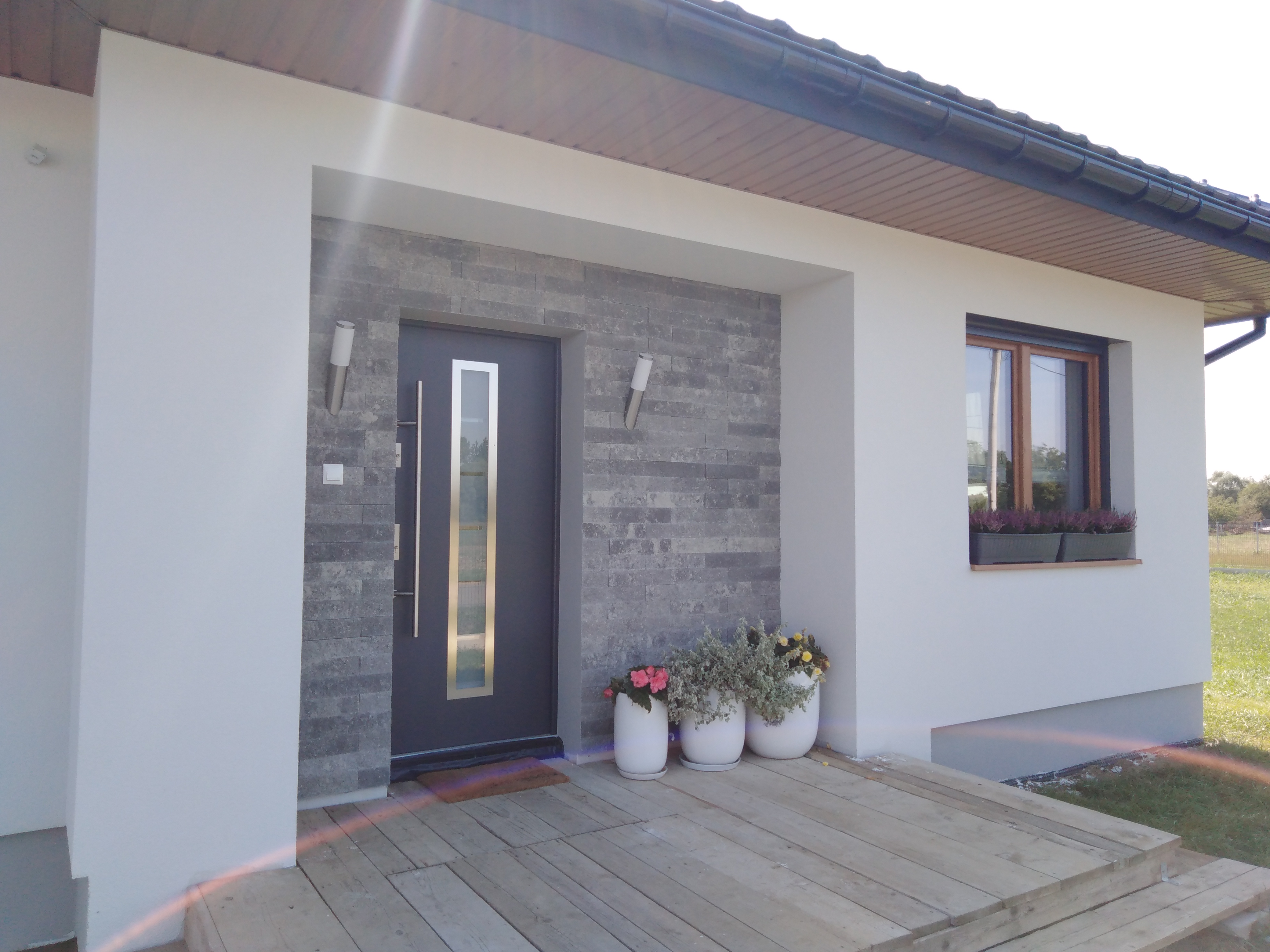 Wykończony fragment elewacji domu jednorodzinnego z szarą, nowoczesną stolarką drzwiową i kamienną okładziną, oświetlony lampami elewacyjnymi i udekorowany kwiatami w donicach.