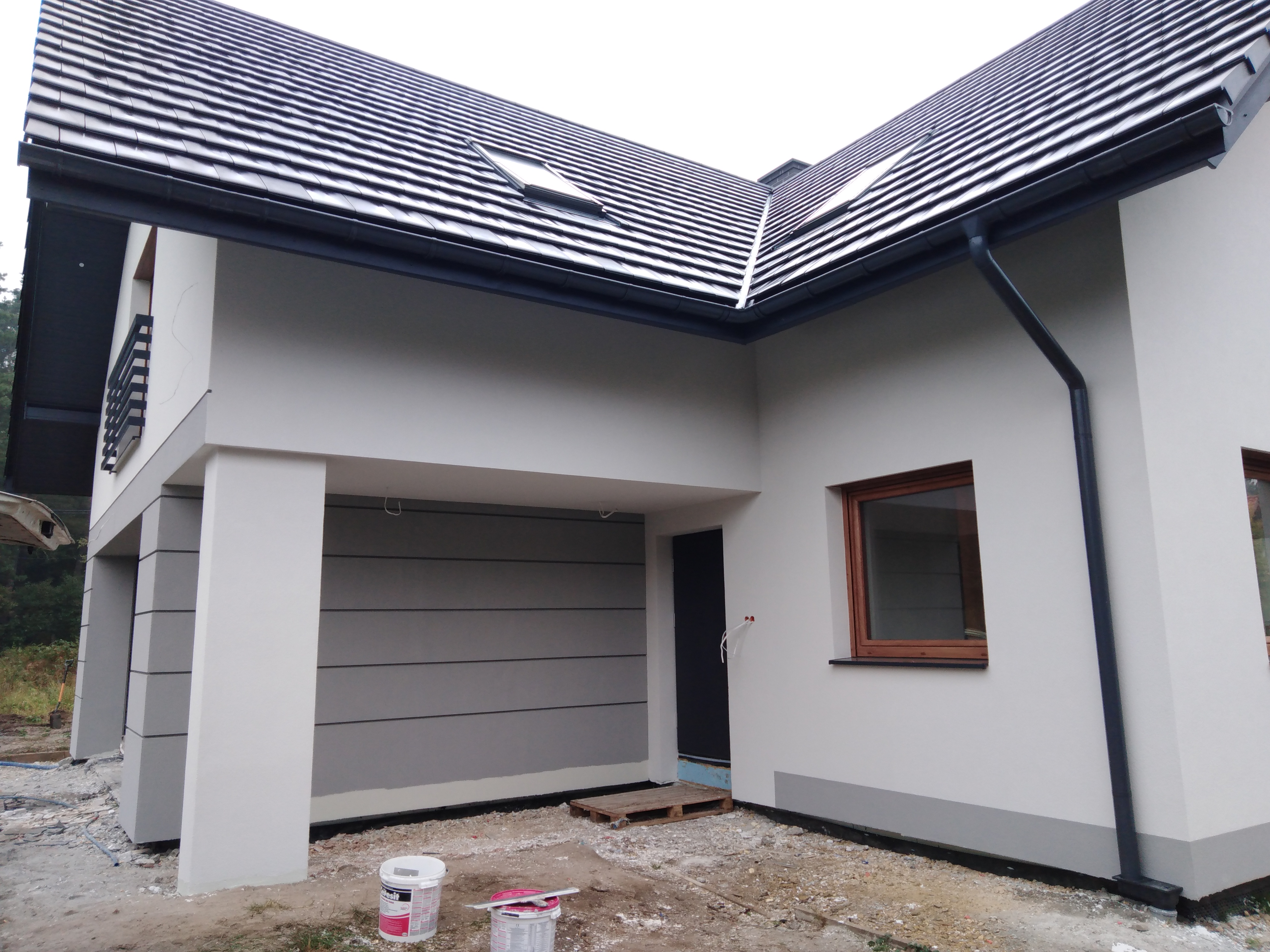 Nowoczesny dom jednorodzinny z szarą elewacją, ciemną dachówką i oknami w drewnianych ramach. Widoczne detale wykończenia, takie jak rynny i obróbki blacharskie w kolorze grafitowym. Na pierwszym...