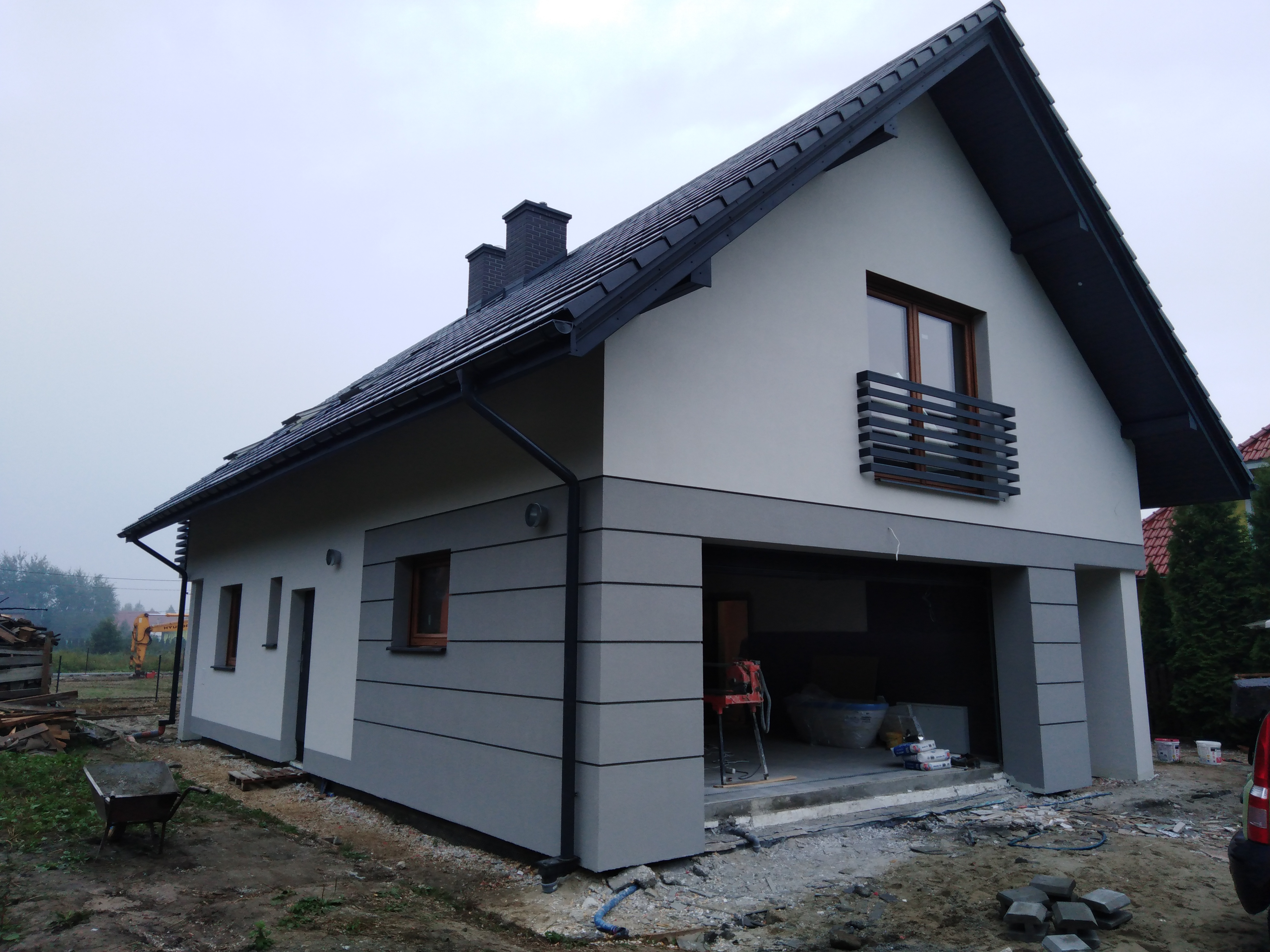 Dwukondygnacyjny dom jednorodzinny z szaro-białą elewacją, czarną dachówką, widocznym garażem w trakcie prac wykończeniowych, z taczką i koparką w tle.