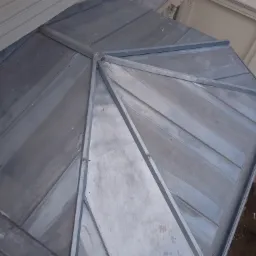 Szary dach z blachy trapezowej z widocznymi łączeniami, fragment budynku z dekoracyjnymi elementami architektonicznymi w tle.