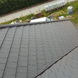 Ukończony dach pokryty szarą dachówką ceramiczną, widok z góry na ułożony wzór, w tle zaparkowany biały bus i materiały budowlane.