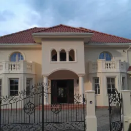 Elegancka, kremowa elewacja domu z czerwonym dachem, widoczna przez ozdobne, czarne, metalowe ogrodzenie z inicjałami, w słoneczny dzień.