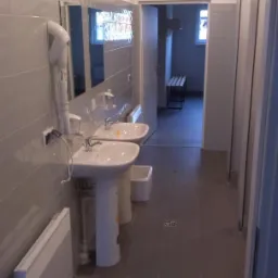 Wnętrze publicznej toalety po remoncie: dwie białe umywalki na postumentach, grzejnik na ścianie, szare płytki, lustro i kabiny WC. Widok na pomieszczenie z ławką.