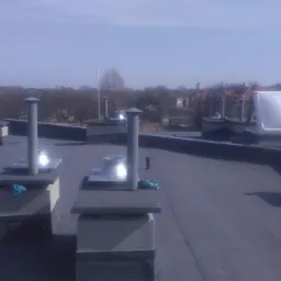 Widok z góry na płaski dach pokryty czarną papą, z kilkoma kominami wentylacyjnymi o różnych kształtach, w tle budynki mieszkalne i drzewa.