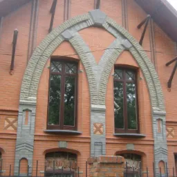 Ceglana elewacja budynku z dwoma oknami w łukowych obramowaniach z szarego kamienia, dekoracyjne elementy z cegły i kamienia, widoczne wsporniki na ścianie.