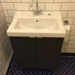 Biała umywalka z szafką w ciemnym kolorze, chromowana bateria i korek, białe płytki na ścianie, ciemnoniebieska podłoga w kratkę.