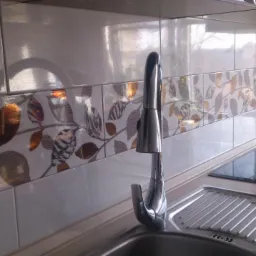 Błyszczące, białe płytki ścienne z dekoracyjnym pasem w motywy liści, chromowana bateria umywalkowa i zlewozmywak w kuchni. Nowoczesne wykończenie.