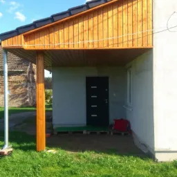 Zadaszenie nad wejściem do budynku z drewnianą konstrukcją i nową elewacją. Widoczna kolumna wspierająca konstrukcję, drzwi wejściowe i fragment trawnika.