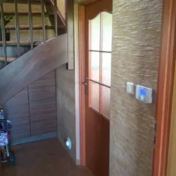 Korytarz z drewnianymi schodami, szafką pod schodami i drzwiami z szybkami. Ściany pokryte dekoracyjną strukturą przypominającą kamień.