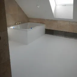 Biała, jednolita podłoga w łazience na poddaszu z oknem dachowym, widoczna wanna obudowana płytkami imitującymi kamień. Podłoga sprawia wrażenie gładkiej i jednolitej powierzchni.