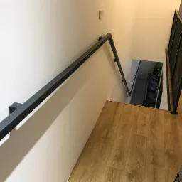 Nowoczesna klatka schodowa z minimalistyczną, metalową balustradą w kolorze grafitowym, o prostych, geometrycznych kształtach, z widoczną perforowaną barierką na podeście i drewnianą podłogą.