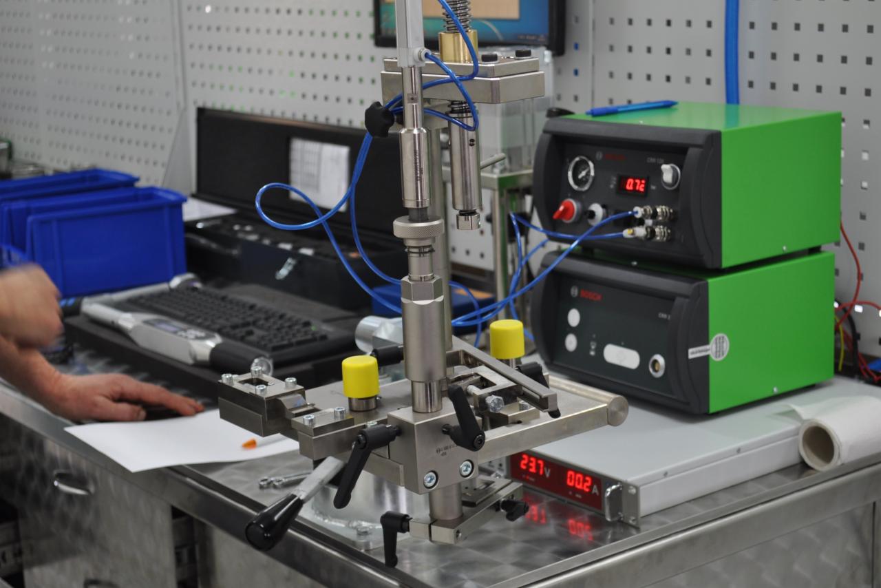 Stół warsztatowy z oprzyrządowaniem do testowania wtryskiwaczy paliwa; widoczne testery firmy Bosch, metalowa konstrukcja z dźwigniami i elementami mocującymi, ręka osoby obsługującej urządzenie.