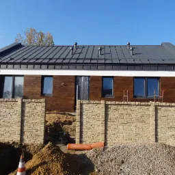 Dom jednorodzinny z drewnianą elewacją i szarą blachą na dachu, ogrodzony murem z piaskowca. Widoczne prace ziemne i materiały budowlane.