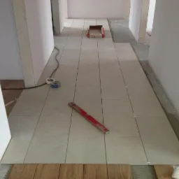 Układanie jasnych, prostokątnych płytek podłogowych w korytarzu. Widoczna poziomica, taśmy malarskie i krzyżyki dystansowe. Częściowo położone drewniane panele.