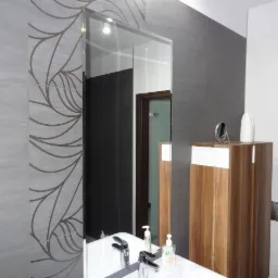 Nowoczesna łazienka z szarymi płytkami zdobionymi motywem roślinnym, dużym lustrem i drewnianą szafką. Widoczne umywalka z baterią i akcesoria łazienkowe.