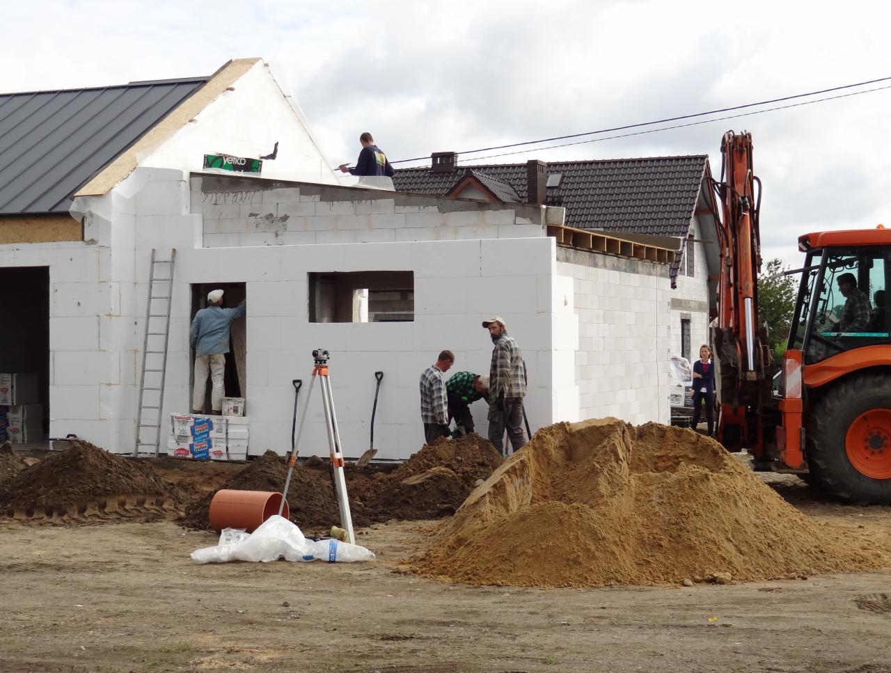 Budowa domu z białych bloczków: ekipa murarzy w akcji, jeden na dachu, drugi maluje ściany wewnątrz, obok stoi pomarańczowa koparka i góra piasku.