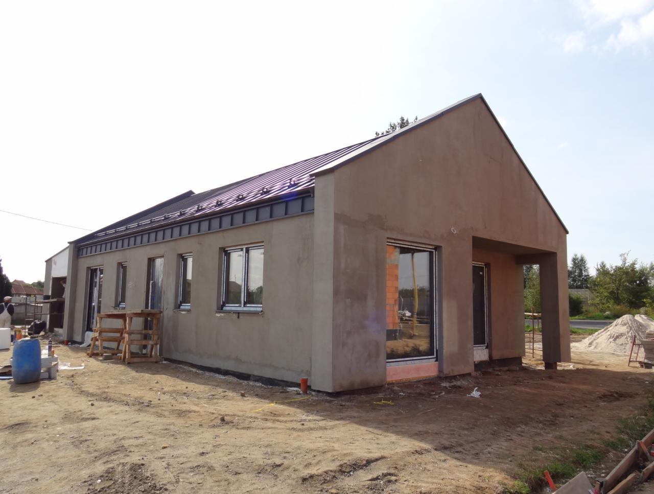 Surowa elewacja domu jednorodzinnego w trakcie prac wykończeniowych. Widoczne okna z czarnymi ramami, błękitne niebo i elementy rusztowania.