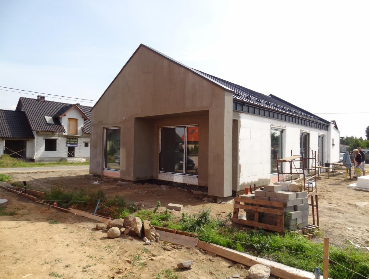 Dom w trakcie budowy z oknami, częściowo otynkowany na beżowo, z czarnym dachem. Widoczne materiały budowlane i sąsiedni dom w trakcie prac elewacyjnych.