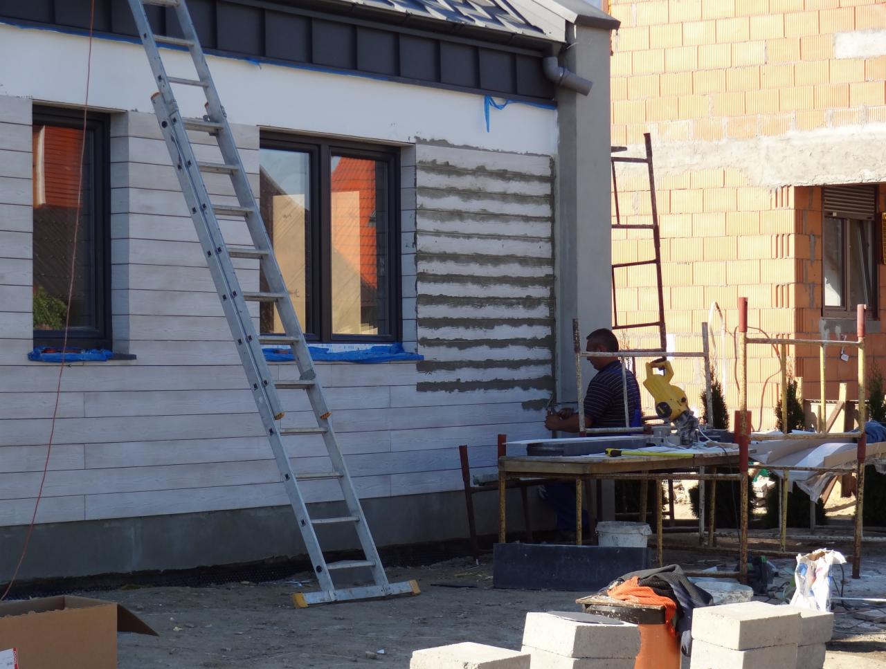 Montaż elewacji z poziomych paneli na budynku. Widoczne rusztowanie, drabina i narzędzia pracy. Fragment elewacji w trakcie montażu, z widocznymi warstwami kleju.