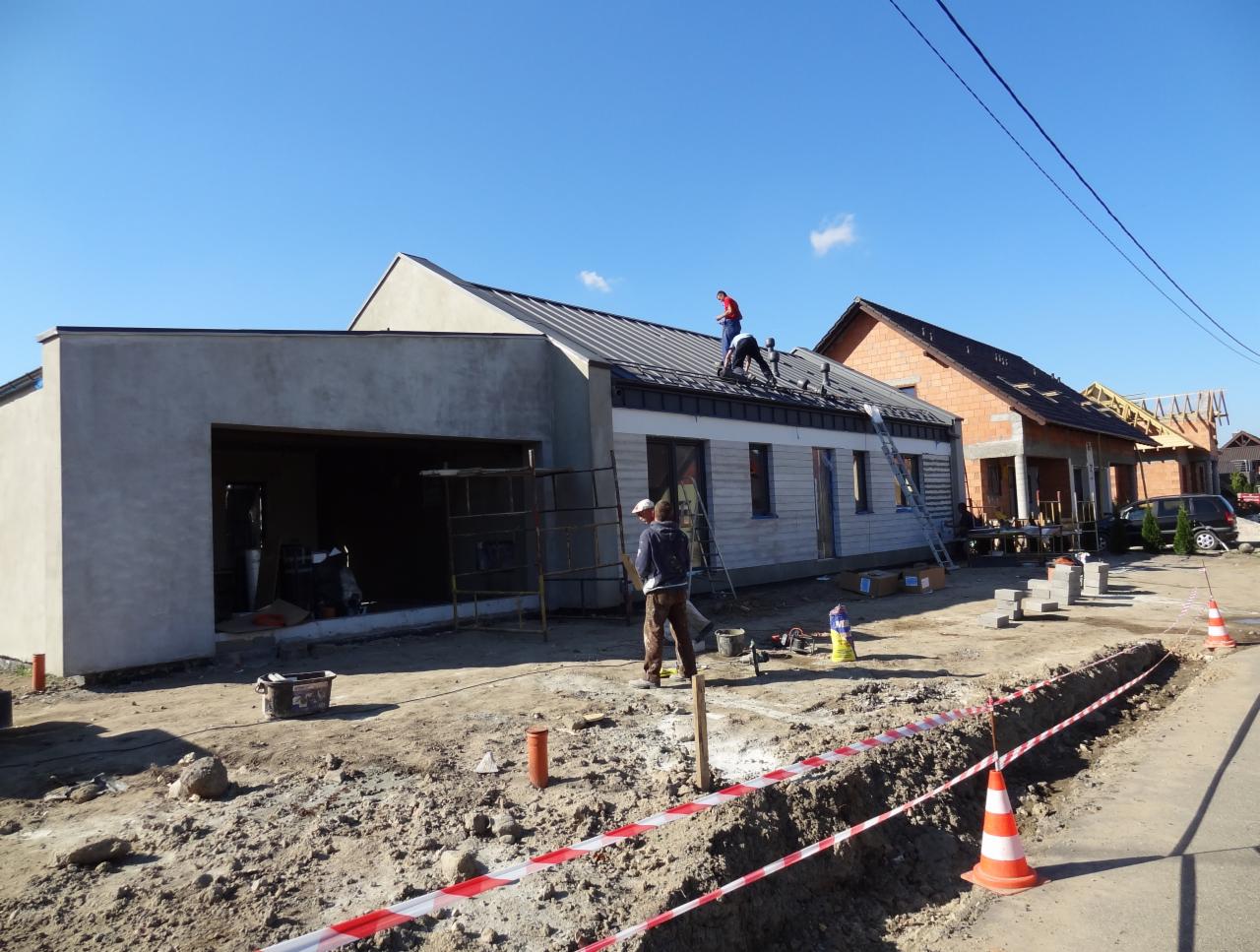 Budowa domu jednorodzinnego: ekipa montuje dach, elewacja w trakcie, widoczne materiały budowlane i narzędzia. Jasne niebo, słoneczny dzień.