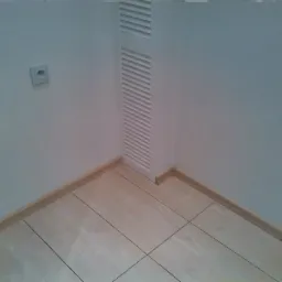 Wykończony fragment łazienki: jasne płytki podłogowe, białe ściany z kratką wentylacyjną i gniazdkiem. Widoczne listwy przypodłogowe i fragment ościeżnicy.