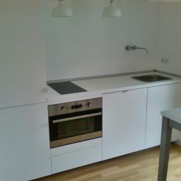 F.U.H Artur LASKOWSKI - Minimalistyczna, biała zabudowa kuchenna z płytą indukcyjną i piekarnikiem, uzupełniona oświetleniem punktowym i drewnianą podłogą w nowoczesnym wnętrzu.