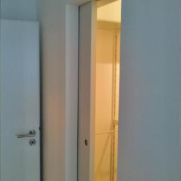 F.U.H Artur LASKOWSKI - Białe, przesuwne drzwi do garderoby w minimalistycznym wnętrzu. Widoczny fragment ściany z drzwiami po lewej stronie. Ciepłe światło wewnątrz garderoby.