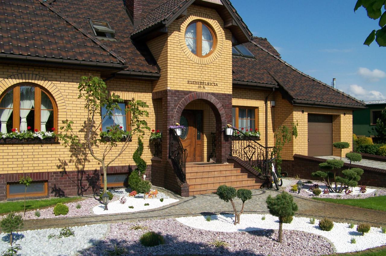 Elewacja domu z żółtej cegły z elementami dekoracyjnymi: okrągłe okno, kwiaty w oknach, ścieżka z białego i różowego kamienia, ozdobne drzewka iglaste.