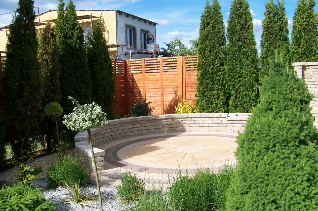 Okrągły plac z kostki, otoczony niskim murem i zielenią. W tle drewniany płot i wysokie tuje. Dekoracyjne drzewko na pierwszym planie.