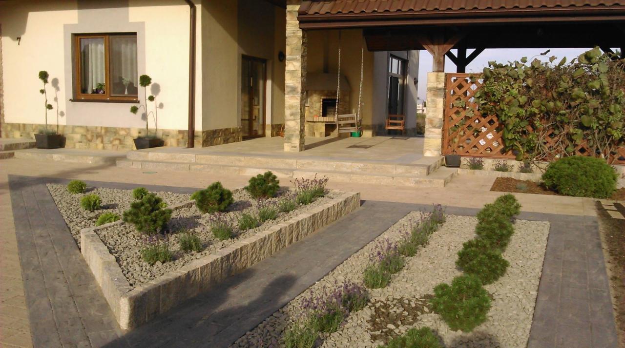 Ogród z niskimi krzewami i kamieniami ozdobnymi, ścieżka z kostki brukowej prowadzi do domu z tarasem i huśtawką. Widoczne rośliny w donicach przy oknie.