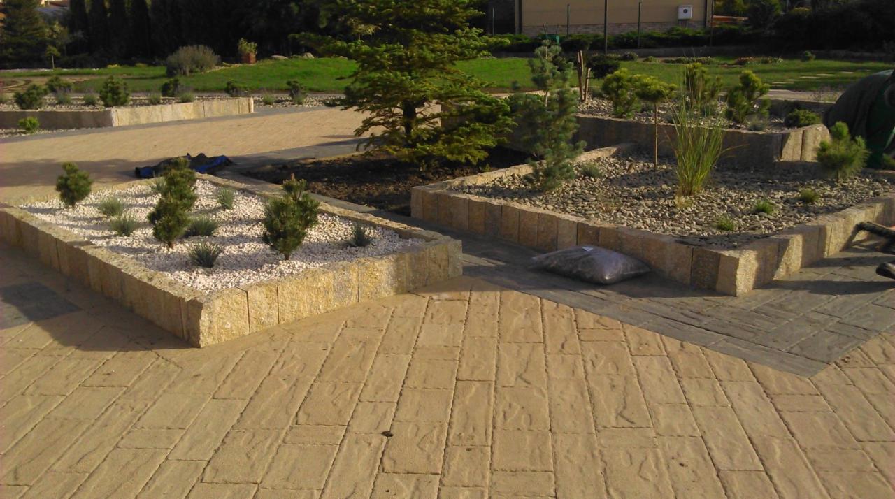 Kwadratowe rabaty z jasnego kamienia, wysypane białym i szarym żwirem, obsadzone iglakami i trawami ozdobnymi. Na pierwszym planie jasna kostka brukowa.