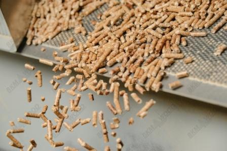 Jasne, drobne granulki na metalowej taśmie, rozsypujące się w dół. Widoczne logo firmy w rogu. Delikatne oświetlenie, neutralne tło.