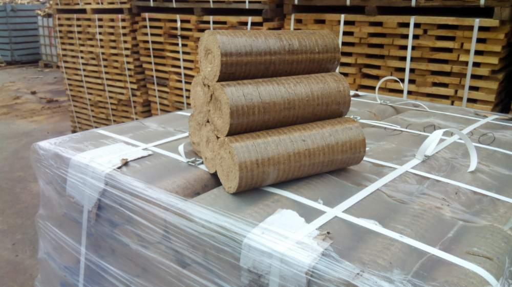 Ułożone na palecie, owiniętej folią, trzy cylindryczne brykiety z trocin na tle drewnianych palet. Widoczne paski mocujące.