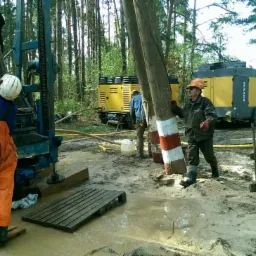 Wiercenie studni w terenie leśnym: operator w kasku obsługuje wiertnicę, obok stoi żółty kompresor. Prace ziemne w błotnistym terenie, widoczne rury i narzędzia.