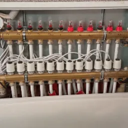 Rozdzielacz ogrzewania podłogowego w szafce z widocznym sterownikiem TECH, zaworami termostatycznymi i rurami doprowadzającymi, zamontowany na tle ściany o strukturze baranka.