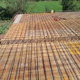 Zbrojenie stropu na deskowaniu z widocznymi prętami zbrojeniowymi ułożonymi w kratę, zabezpieczonymi drewnianymi deskami i elementami szalunku, z widokiem na zieloną trawę w tle.